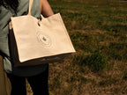 A person outdoors carrying a large natural jute and cotton tote bag with a circular green logo embroidered on the front. The background is a grassy field.