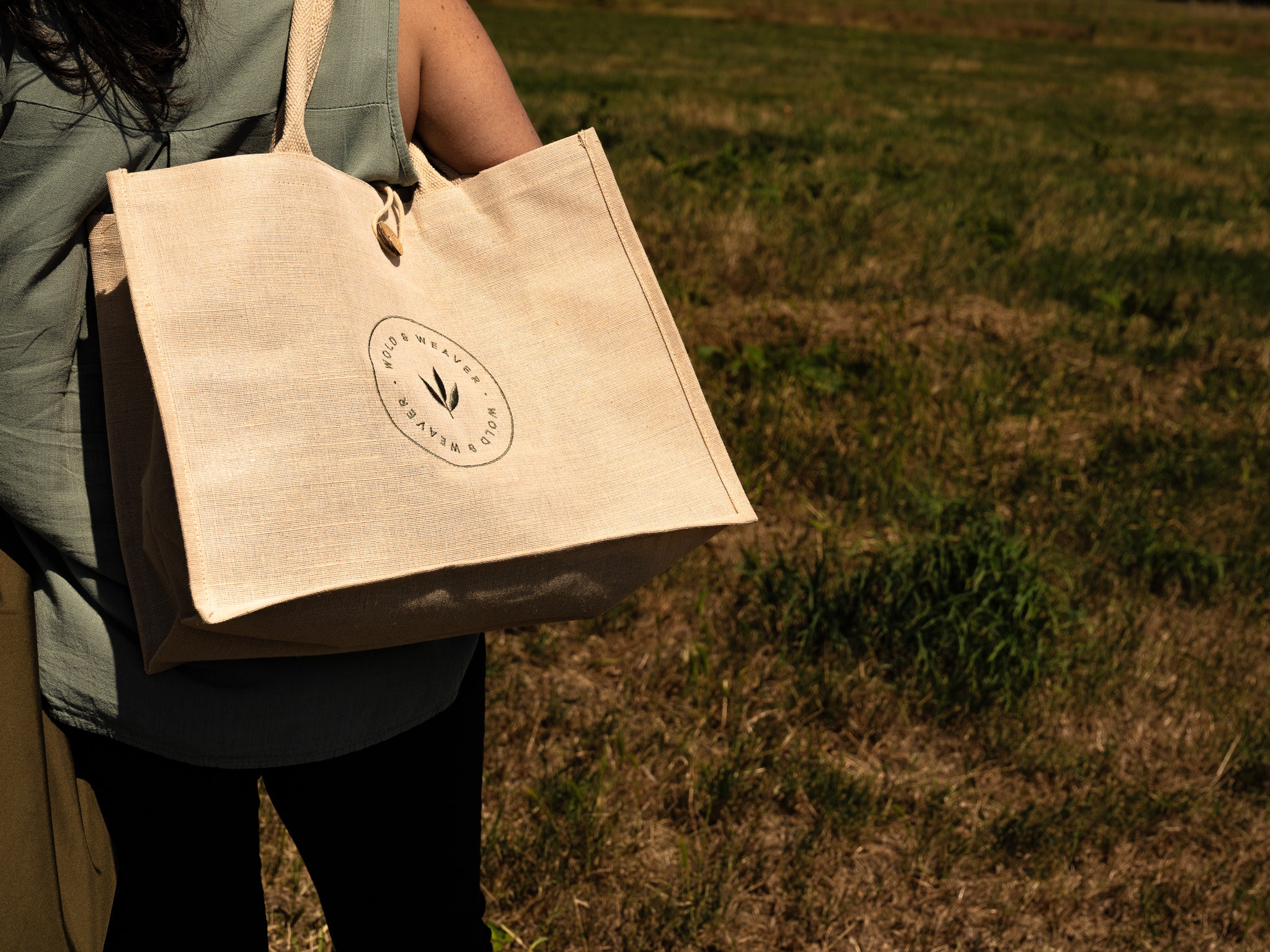 A person outdoors carrying a large natural jute and cotton tote bag with a circular green logo embroidered on the front. The background is a grassy field.