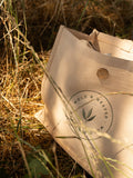 A beige canvas tote bag with a circular 'Wold & Weaver' logo embroidered on the front, resting on grass outdoors in the countryside.