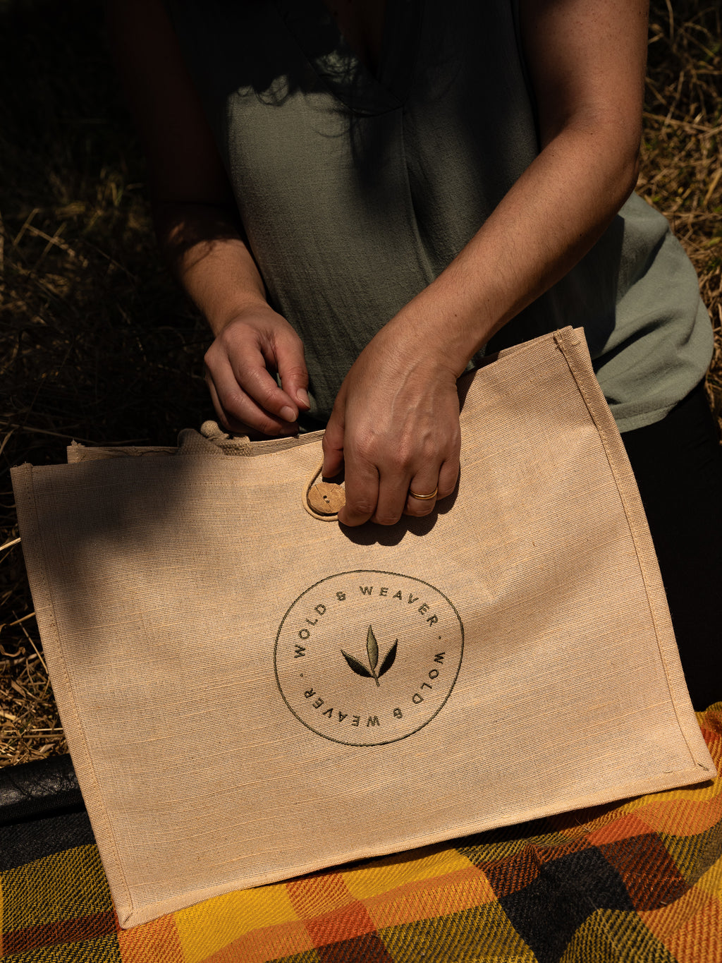 A person outdoors holds a large natural jute and cotton shopping bag with a circular 'Wold & Weaver' logo embroidered on the front. The scene is lit by warm, cosy sunlight with a checked picnic blanket underneath.