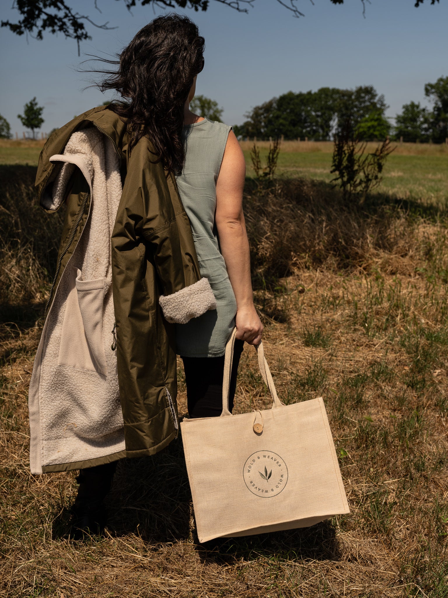 A woman standing outdoors in a grassy field, seen from behind, carrying an olive green hooded jacket with a cream fleece lining and carrying a natural cream jute and cotton shopping bag with a circular logo that reads 'Wold & Weaver'.