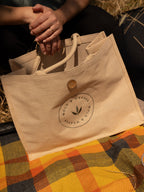 A beige natural jute and cotton tote bag with cotton handles and a circular 'Wold & Weaver' logo, held by a person sitting on a checked picnic blanket outdoors.