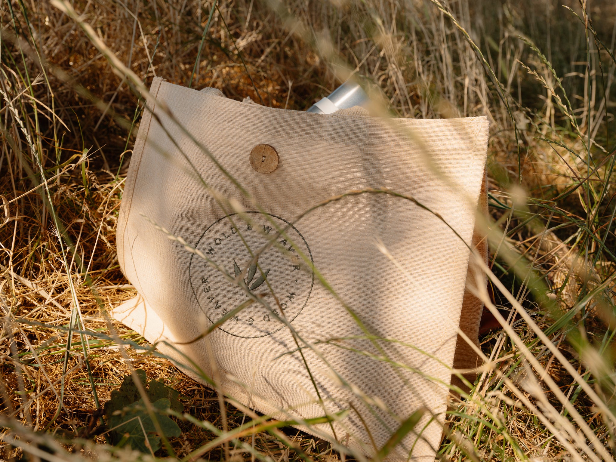 A natural beige jute and cotton picnic bag with a circular 'Wold & Weaver' logo embroidered on the front, resting on grass in an outdoor countryside setting.