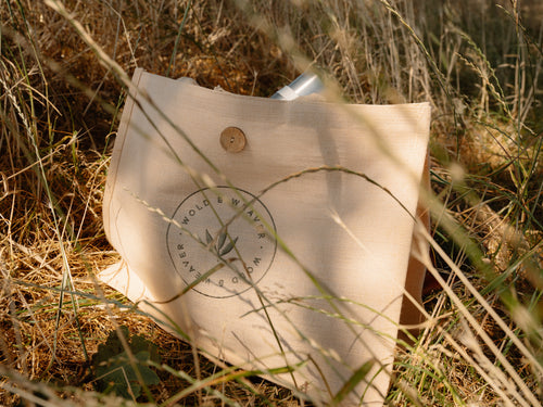 A natural beige jute and cotton picnic bag with a circular 'Wold & Weaver' logo embroidered on the front, resting on grass in an outdoor countryside setting.
