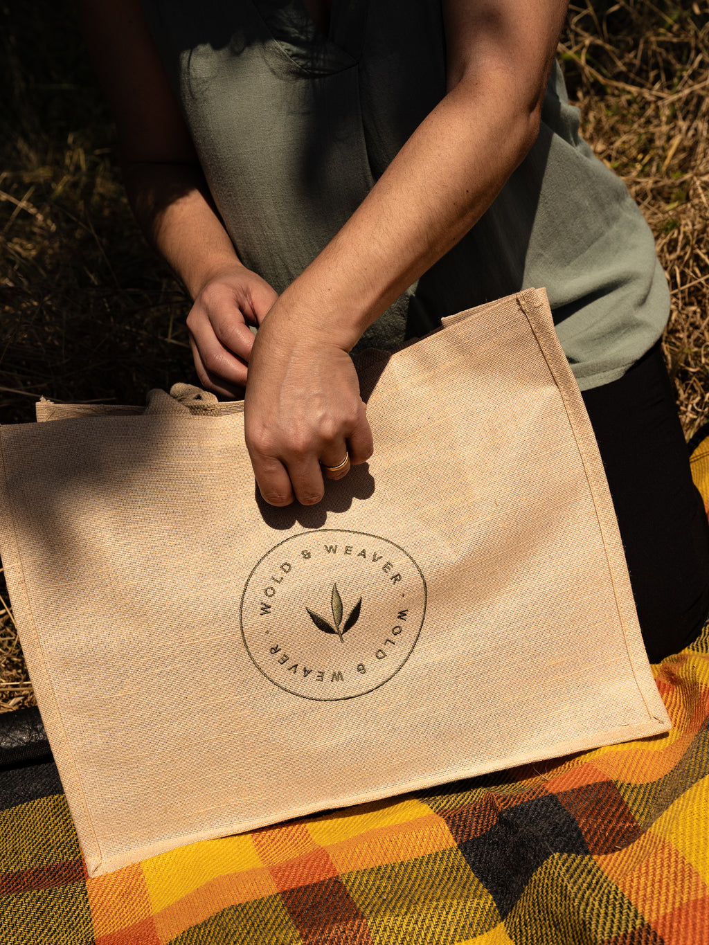 A person outdoors holds a natural jute and cotton picnic bag printed with a circular 'Wold & Weaver' logo. The bag rests on a yellow, orange, and black plaid picnic blanket.