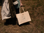 A woman outdoors on grass holding a natural jute and cotton shopping bag with a circular logo embroidered on the front. A country robe is visible on the left.