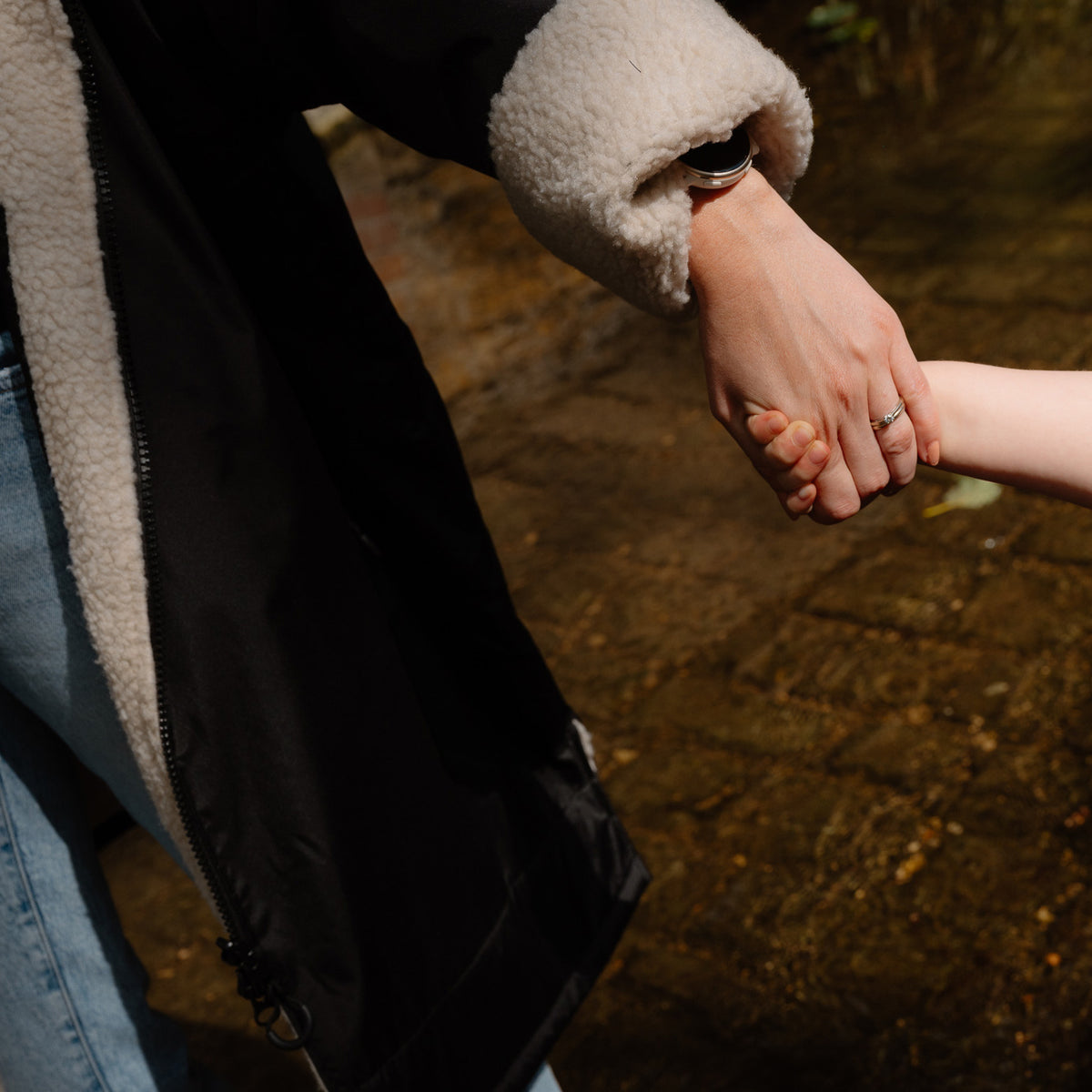 A close-up lifestyle shot of a person in a black waterproof jacket with a cream fleece lining holding hands with a child outdoors on a stone path.
