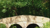Woman wearing waterproof country robe walks slowly over a bridge in a Cotswolds village surrounded by green foliage and countryside.