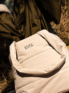 A close-up of a light beige stone quilted puffer bag with handles and with the brand name 'Wold & Weaver' embroidered in dark green on the front, set against a background of an olive green parka jacket and dry grass.
