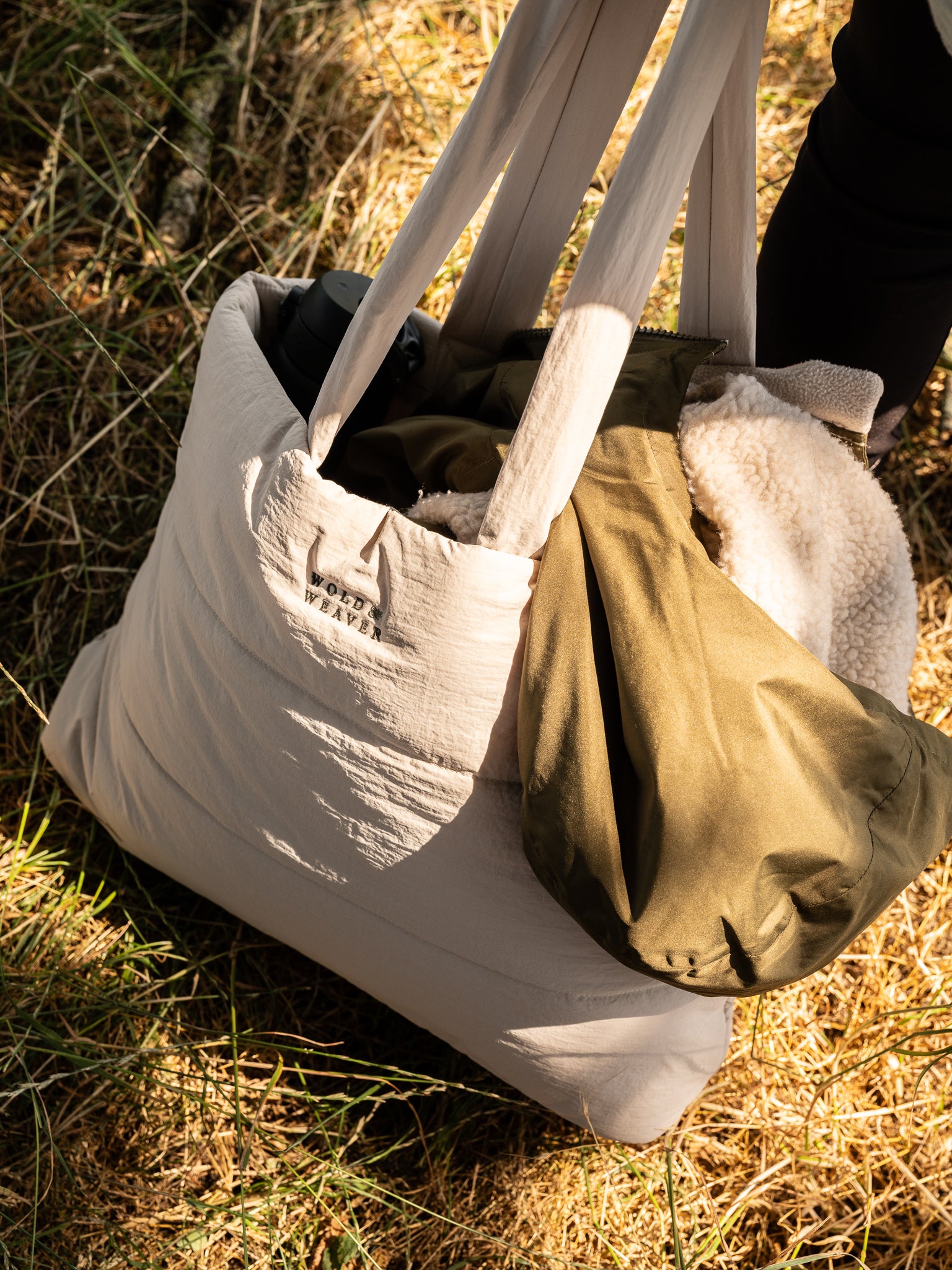 A large 100 percent recycled stone coloured quilted tote bag with comfortable shoulder straps, shown outdoors on grass with an olive green hooded coat tucked inside. The brand name 'Wold & Weaver' is visible in a small woven logo on the front.