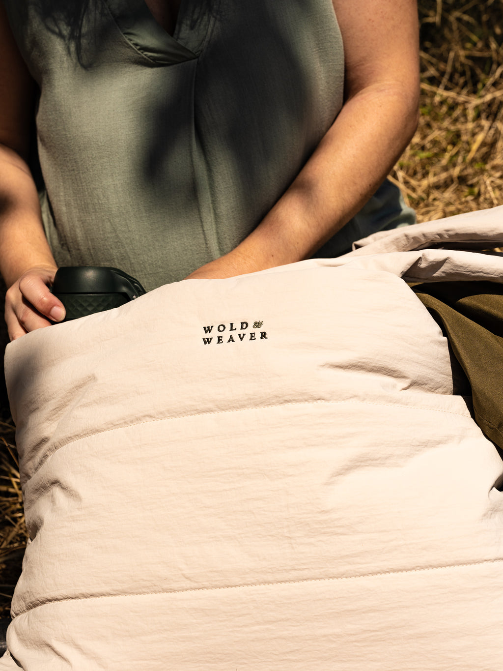 A close-up of a person outdoors holding a light beige quilted puffer bag with the brand name 'WOLD & WEAVER' embroidered in dark green. The person is wearing a muted sage green sleeveless top, and a grass field is visible in the background.