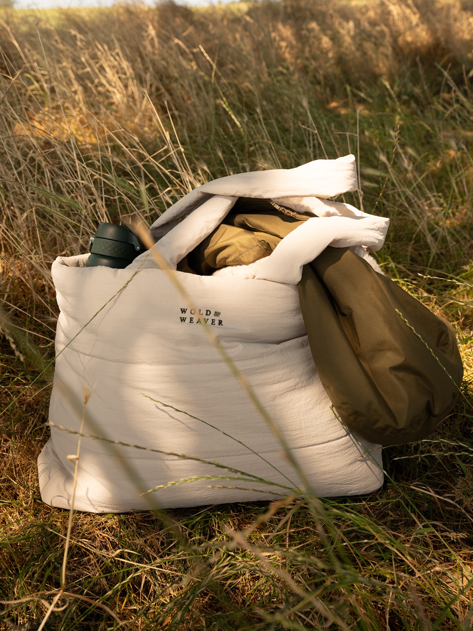 A large off-white quilted tote bag sits on grass outdoors, with a dark green bottle tucked into the top opening and an olive green jacket hanging from the side. The bag has the text 'Wold & Weaver' embroidered near the top front.