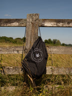 A black drawstring bag for a waterproof country robe with a white circular 'Wold & Weaver' logo hanging from a weathered wooden fence in an outdoor grassy field under a blue sky.