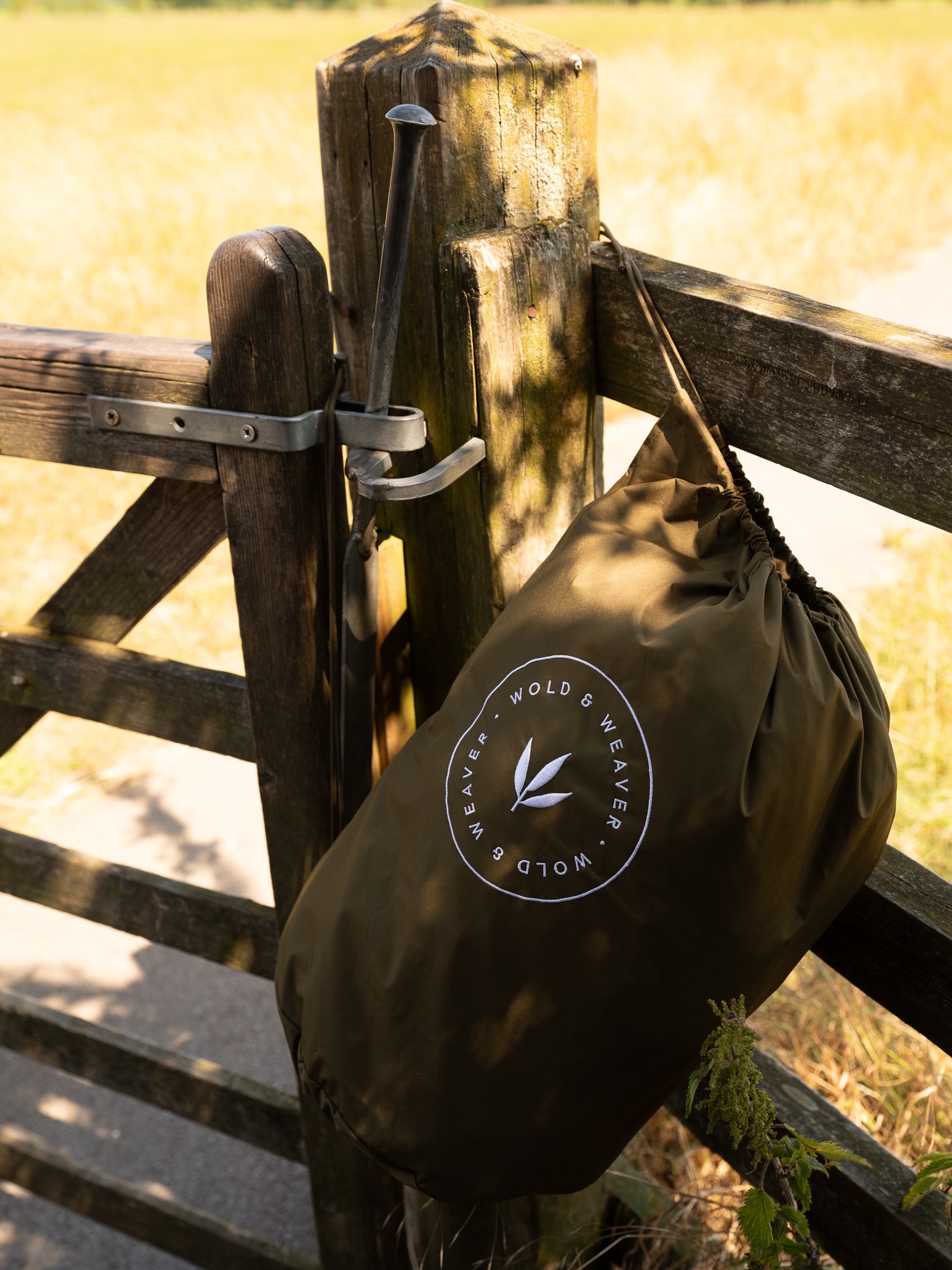 An olive green drawstring bag for the waterproof country robe, with a circular white logo hanging from a wooden gate in an outdoor grassy setting. The logo reads 'Wold & Weaver'.