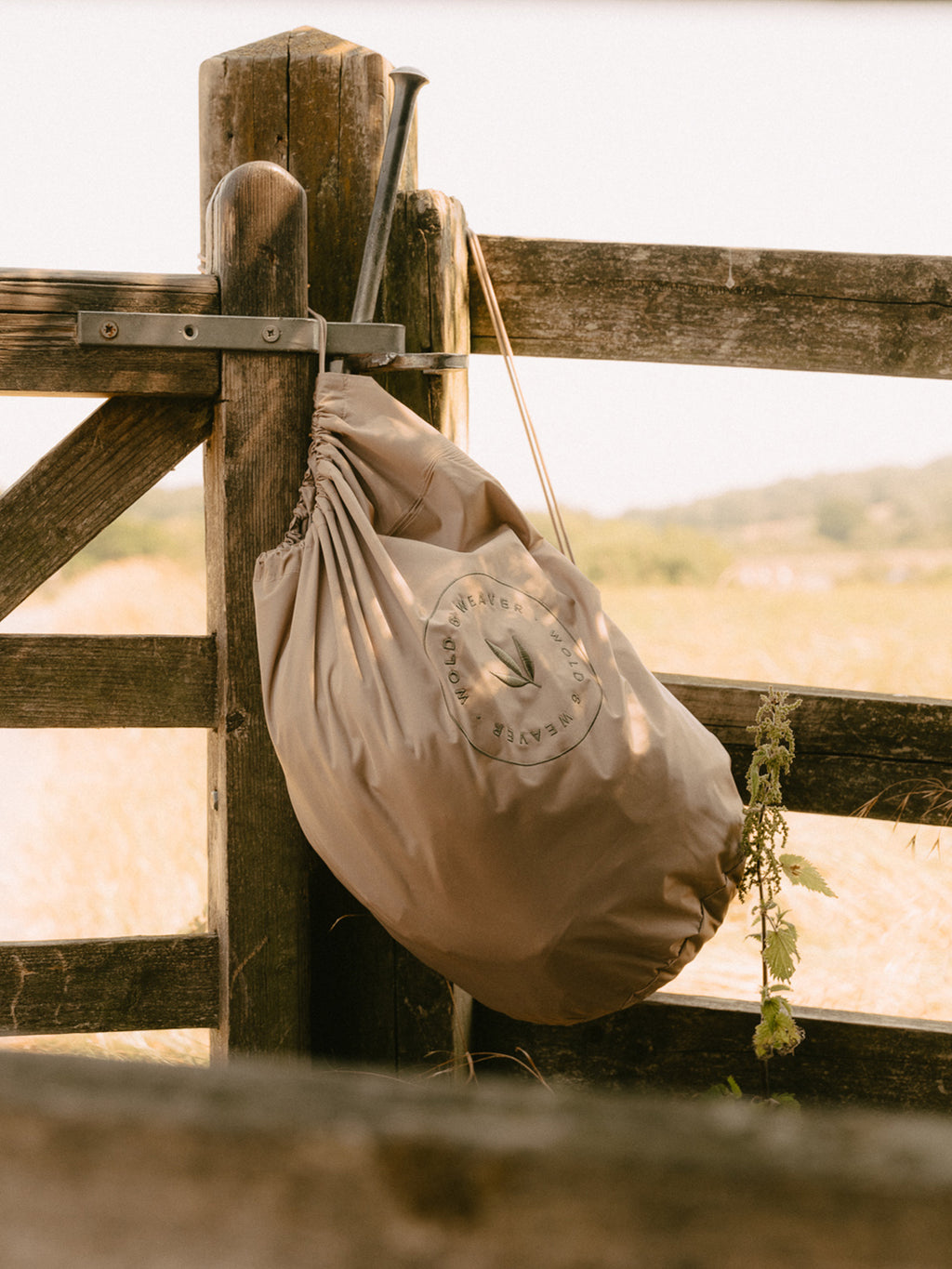 A beige drawstring bag containing the waterproof robe with a circular logo hanging from a rustic wooden fence gate in an outdoor field setting.