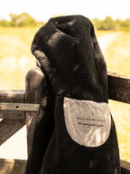 A black fleece-lined hooded coat draped over a rustic wooden gate, with a light beige brand patch reading 'WOLD & WEAVER The countryside is yours' visible on the inside.