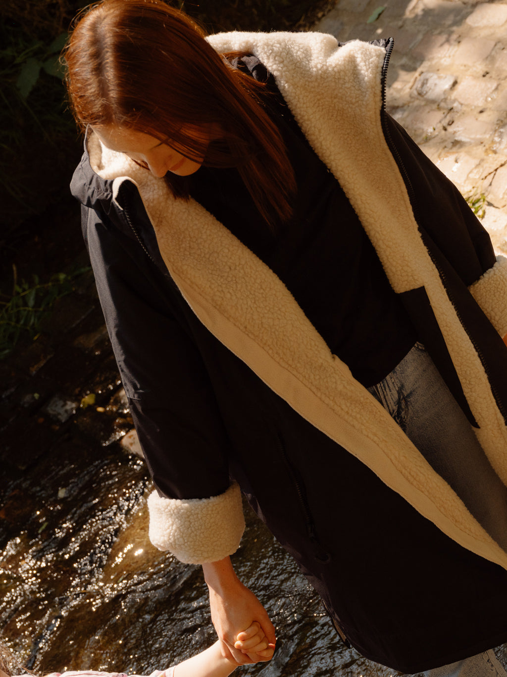A woman wearing a black waterproof country robe coat with a thick cream sherpa lining stands in shallow water outdoors, holding a child's hand in the water.