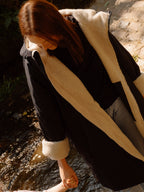 A woman wearing a black waterproof country robe coat with a thick cream sherpa lining stands in shallow water outdoors, holding a child's hand in the water.