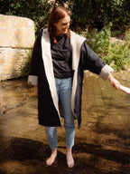 A woman standing barefoot in shallow water outdoors, wearing a black waterproof coat with a thick cream shearling-style lining and light blue jeans.