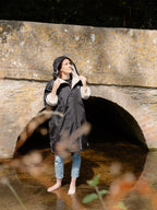 A woman wearing a black hooded raincoat with a cream fleece lining stands barefoot under a stone bridge in shallow water in the Cotswolds.
