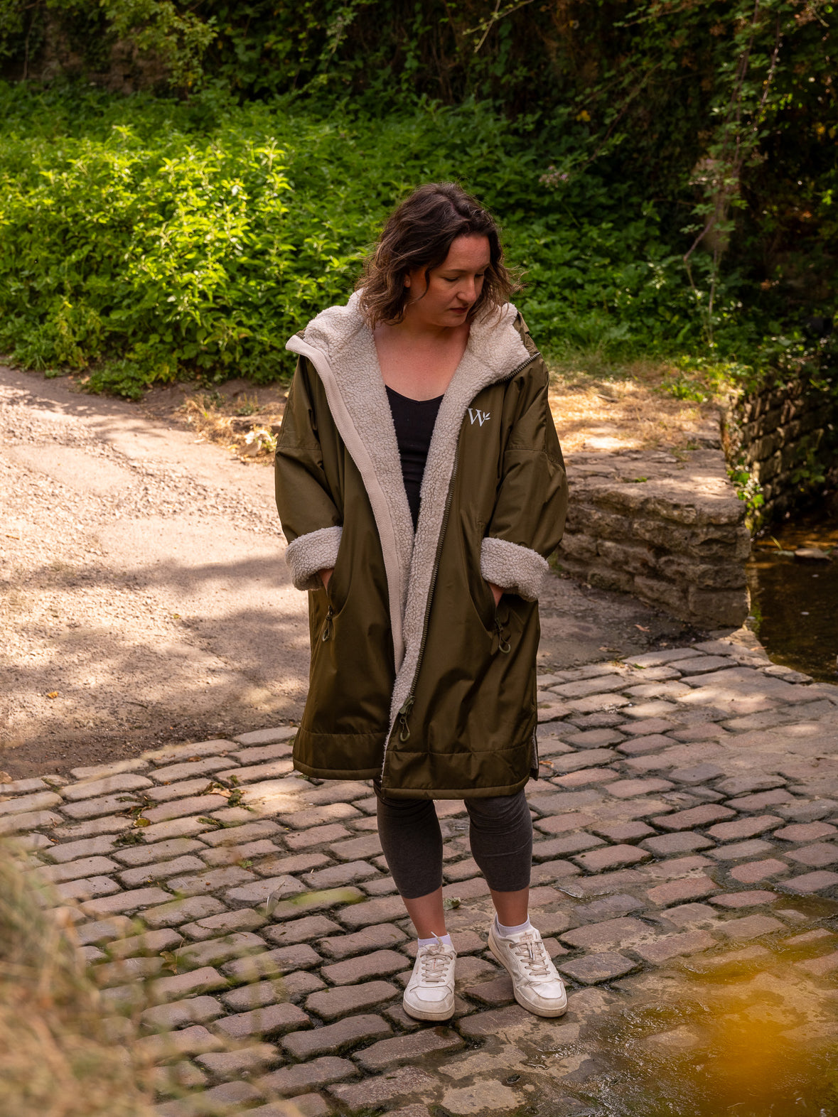 A woman wearing a long olive green country robe with a natural cream sherpa lining, standing on a cobblestone path outdoors in the Cotswolds with green foliage in the background.