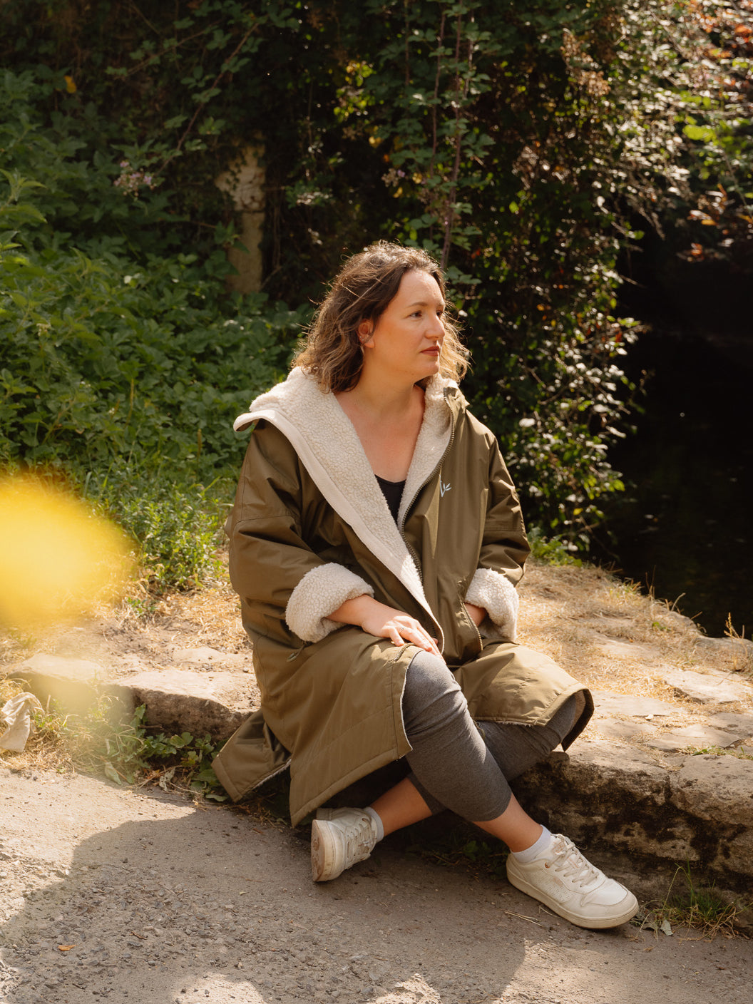 A woman sits on a stone ledge in a Cotswolds village wearing a long olive green waterproof and windproof country robe with a cream sherpa fleece lining, grey leggings, and white trainers, with dense green foliage in the background.