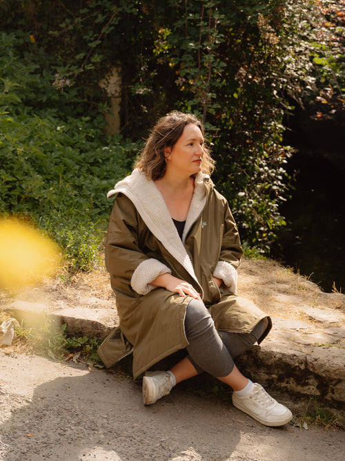 A woman sits on a stone ledge in a Cotswolds village wearing a long olive green waterproof and windproof country robe with a cream sherpa fleece lining, grey leggings, and white trainers, with dense green foliage in the background.