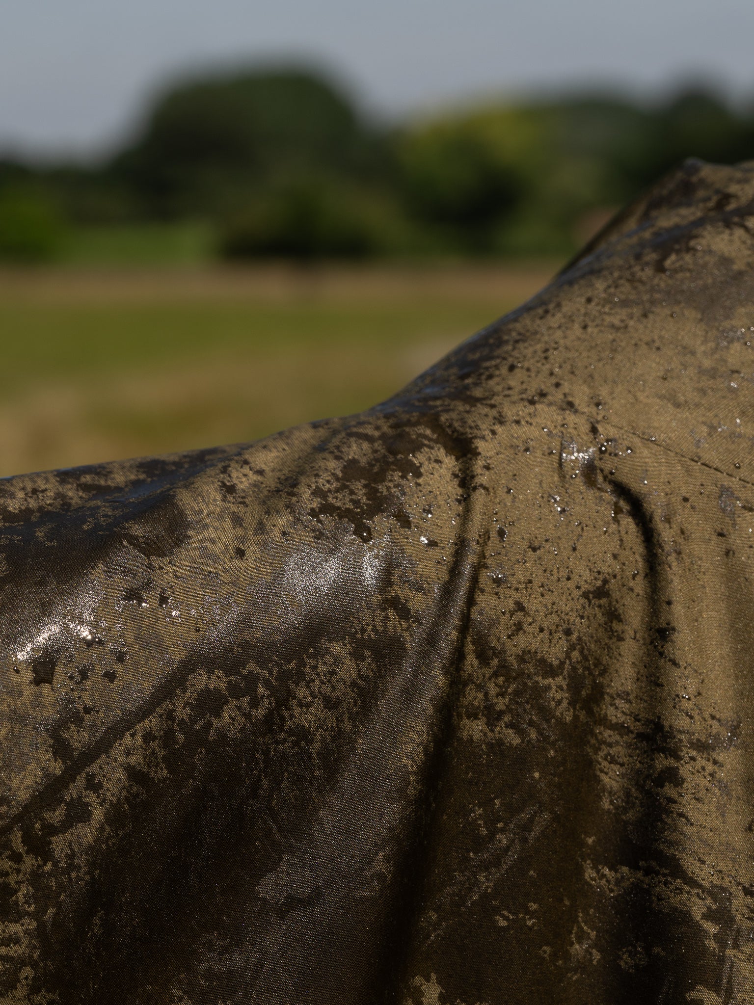 Close-up of a green waterproof country robe coat surface with wet rain water, set against a blurred Cotswolds countryside background of green grass and trees.