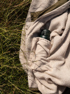 A close-up outdoor shot of a natural cream fleece pocket holding a dark green water bottle, with tall grass visible around it.