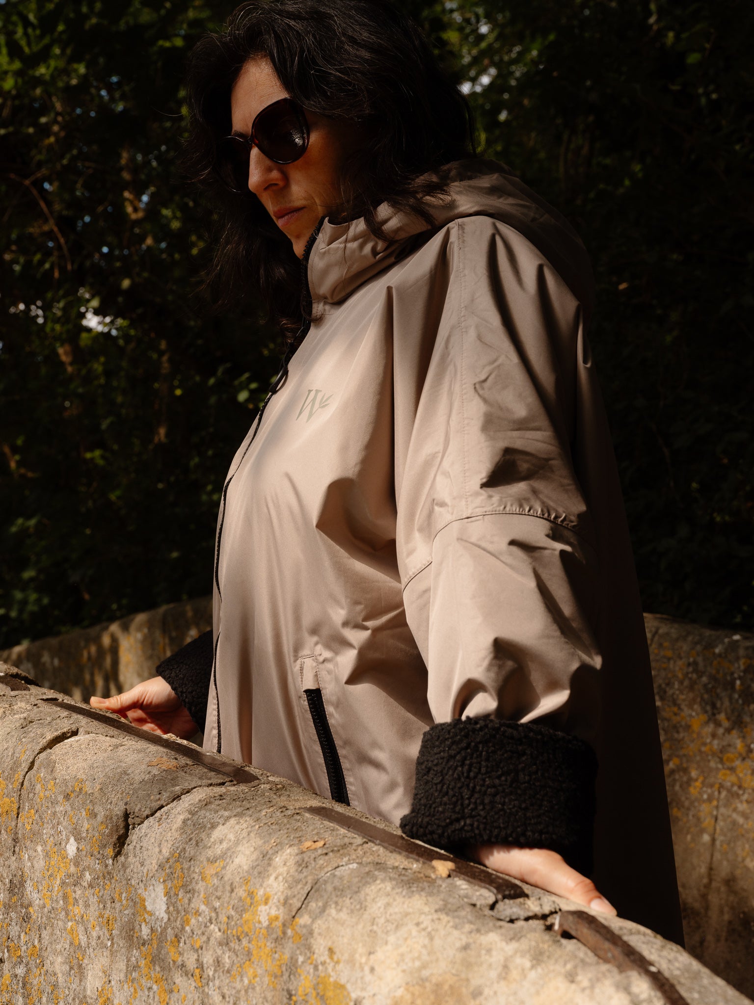 A woman wearing a beige hooded rain jacket with black fleece-lined cuffs and sunglasses, leaning on a stone wall outdoors against a dark, shadowed background.