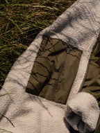 Close-up of a natural fleece-lined jacket laid outdoors on grass, showing a light cream sherpa lining and an olive green waterproof panel with a zipper pocket.