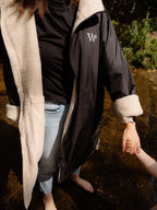 A person wearing a black waterproof and windproof country robe coat with a cream sherpa fleece lining and a white embroidered logo emblem on the chest, standing barefoot in a shallow river stream outdoors.