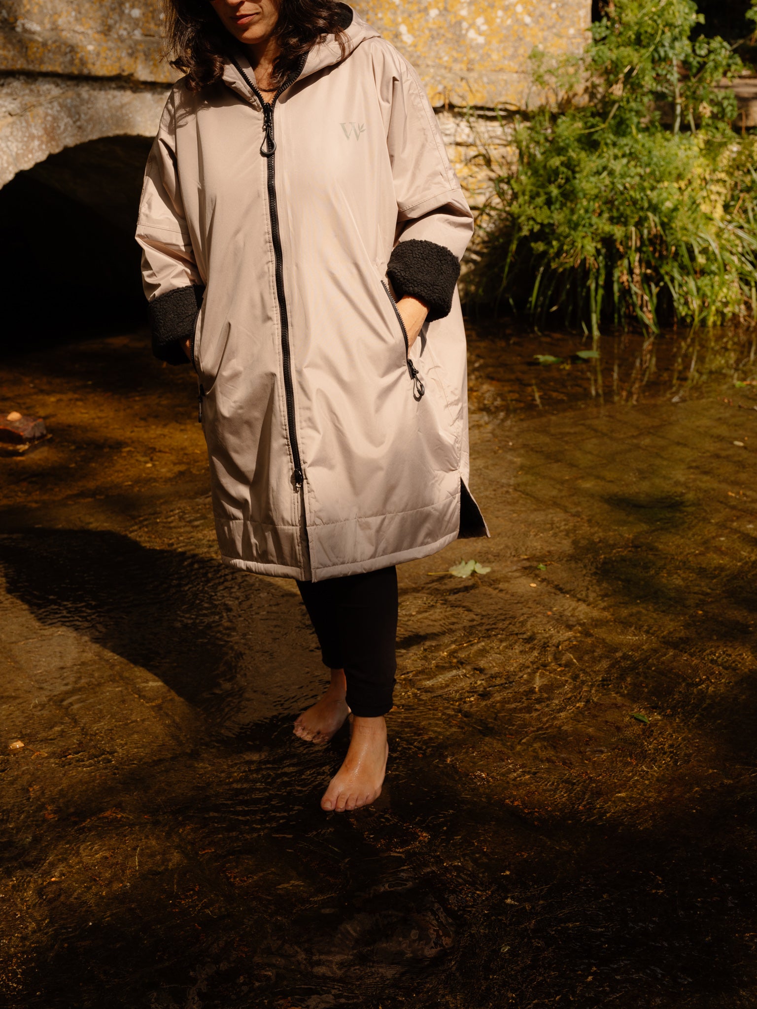 A woman wearing a light beige stone-coloured hooded raincoat with black adjustable cuffs and a black front zipper, standing barefoot in shallow water outdoors near a Cotswolds stone wall and greenery.