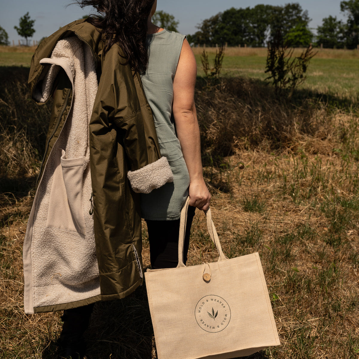 A woman standing outdoors in a field holding a beige natural jute picnic shopping bag with a circular logo that reads 'Wold & Weaver'. The person is also wearing an olive green waterproof hooded jacket with a cream sherpa lining over a sleeveless sage green top.